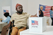 © pressmaster - African American young man with disability sitting in wheelchair and putting ballot paper into box with USA flag standing on desk