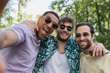 Wall Mural - excited interracial friends in sunglasses hugging in summer park.