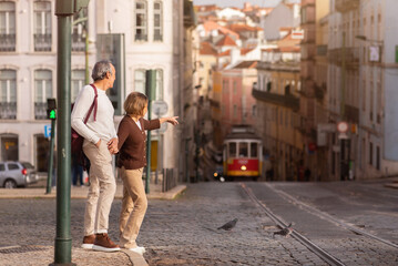 senior tourists couple walking in lisbon, pointing finger at tram