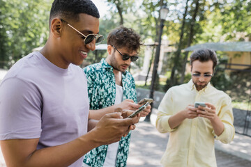 Wall Mural - happy and stylish african american man using smartphone near blurred friends in city park.