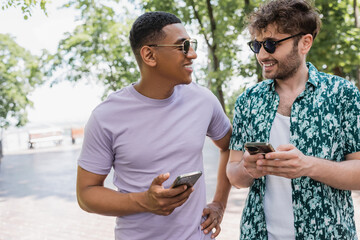 Wall Mural - joyful african american man in sunglasses holding mobile phone and talking to trendy friend in urban