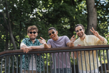 Wall Mural - positive interracial friends in sunglasses gesturing near railing in summer park.