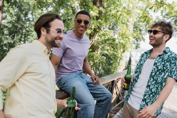 Wall Mural - smiling interracial men in sunglasses holding bottles of beer in park in summer.