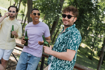 Wall Mural - cheerful man in sunglasses standing near interracial friends with beer in summer park.