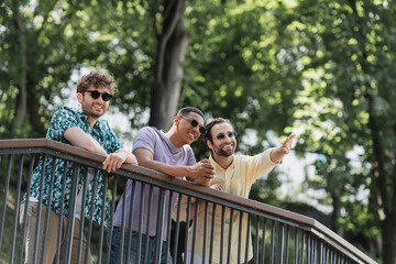 Wall Mural - smiling man pointing with hand near interracial friends in sunglasses in summer park.
