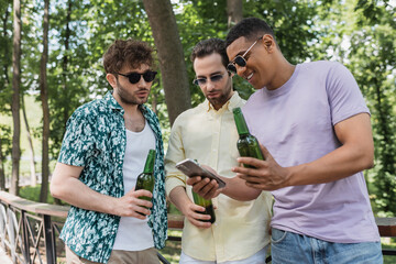 Wall Mural - cheerful african american man in sunglasses showing mobile phone to trendy friends holding beer in c