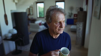 One senior man standing by window drinking tea or coffee in the morning. A pensive older person in 70s looking out in contemplation