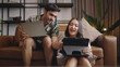© saksit - Young couple using tablet and laptop together on sofa in living room at home