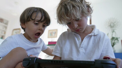 Wall Mural - Two small boys looking at video game screen tablet. Younger brother watching older sibling play games on joystick console. Kids playing with technological device