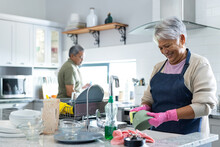 Smiling Biracial Senior Woman Cleaning Bowl In Sink With Husband Standing In Background In Kitchen