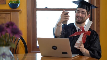 Wall Mural - Young man in graduation costume in front of laptop in a home, taking a selfie.