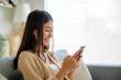 © Monster Ztudio - Happy young asian woman relax on comfortable couch at home texting messaging on smartphone, smiling girl use cellphone chatting, browse wireless internet on gadget, shopping online from home