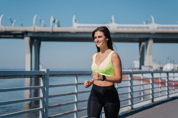 Outdoor shoot of young brunette tanned woman running among embankment, smiling broadly with happy face expression and fluttering hair. Gorgeous hispanic girl in sportswear against bay and large bridge