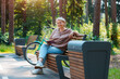 Dreamy modern old lady relaxing in city park. Pensive senior grey haired woman in casual sitting wooden bench outdoors.