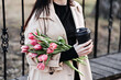 Spring outdoor portrait of happy woman with tulips bouquet and coffee to go on spring city street. Happy young woman holding bouquet of pink flowers