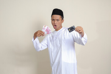 Portrait of attractive Asian muslim man in white shirt with skullcap holding one hundred thousand rupiah and presenting credit card. Isolated image on gray background