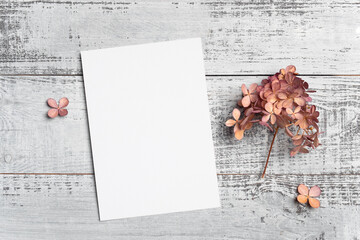 Invitation or greeting card mockup with flowers over wooden background, top view, copy space