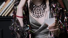 Young woman in colorful glitter blouse transfers wineglass from hand to hand. Lady in silver dress wears nice necklace and red bracelet