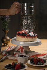 Poster - Chef's hand adding powdered sugar on a delicious cake with berries on a white ceramic stand