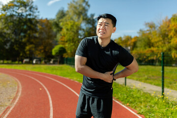 Asian sportsman has severe pain in back and abdominal muscles, man peed training in stadium on sunny day massaging side of back, athlete on jogging pulled muscle.