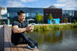 © Liubomir - Asian sportsman resting after training sitting on a bench in the park near the lake and using the phone, the man is smiling and happy about his daily training outdoors on a sunny day.