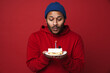 © Drobot Dean - Indian man holding birthday cake and blowing out candle while standing isolated over red wall