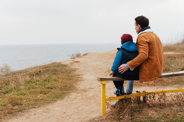 Wall Mural - Back view of father and son enjoying sea view while sitting on bench at beach