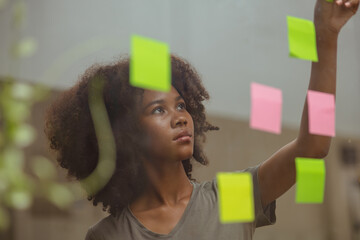 Young African American stylish woman working on project plan using sticky papers notes on glass wall, people meeting to share idea, positive thinking workshop and business design planning concepts.