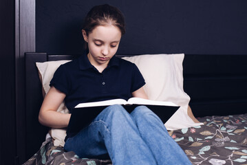 Wall Mural - A teenage girl is reading a book while sitting on the bed. A child reads the Bible. Prayer