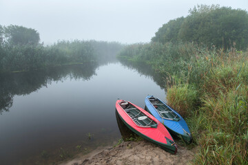 Two multi-colored kayaks on the river bank in the morning fog among the river grass.