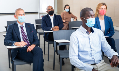 Wall Mural - International group of business people wearing protective face masks listening to presentation in conference room. Concept of precautions and social distancing in COVID 19 pandemic..