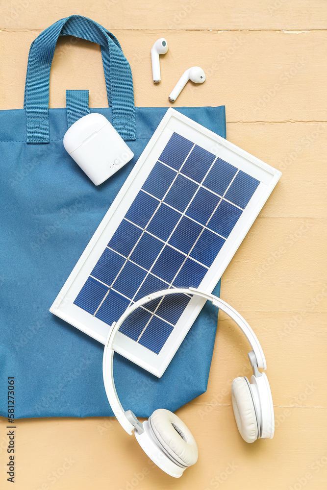 Composition with portable solar panel, wireless earphones and bag on beige wooden background