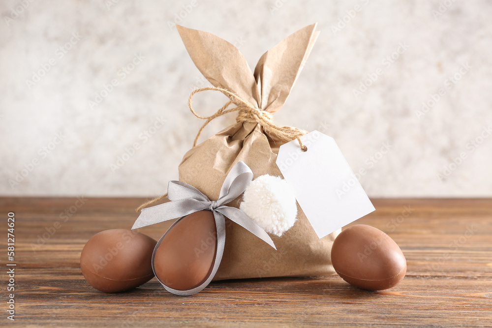 Bunny gift bag with blank tag and Easter eggs on wooden table against grunge wall