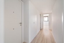 Narrow White Corridor With Doors And Entrances To Different Rooms And Premises And Resting On A Room With A Window On The Sunny Side. Concept Of Minimalist Design In A New Apartment