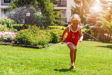 Having Fun With Water Garden Sprinklers In Beautiful Garden With Flowers Bed. Child Running Barefoot On Nature Background. Happy Summer Holidays