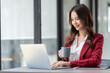 © crizzystudio - Beautiful Asian businesswoman holding a cup of coffee relax Take breaks and work hard on financial business matters at the office and have fun. Smiling happy working with the laptop.