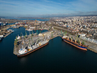 Aerial view of a bulk carrier ship loaded with grain is docked at a busy port, with workers and machinery seen in the background.