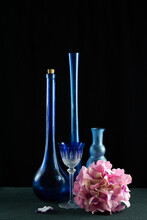 Deux Bouteilles Allongées, Un Verre En Crystal Taillé, Un Vase Bubble Vide Une Fleur D'hortensia Rose  Posée Sur Fond Noir