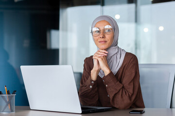 Wall Mural - Portrait of successful and confident Muslim woman in hijab. Director, owner, business woman sitting in the office at a desk with a laptop.