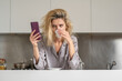 © Volodymyr - Upset woman reading message or bad new at smartphone in the kitchen. Sad woman wearing sexy pajama drink coffee in home kitchen.