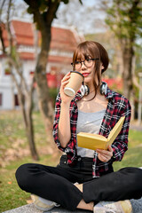 Wall Mural - Beautiful young Asian female college student is sipping coffee and reading a book on a bench