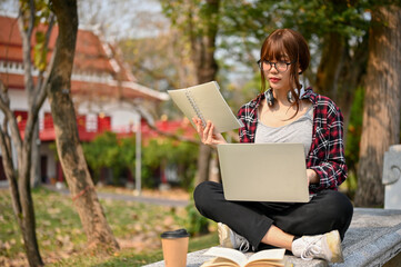 Wall Mural - Attractive Asian female college student using laptop on a bench in campus park.