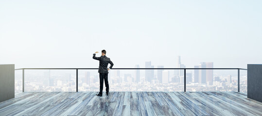 Poster - Back view of young businessman looking into the distance on rooftop with bright daylight sky and wide city view. Success, future and vision concept.