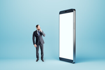 Canvas Print - Thoughtful young businessman looking at big empty white smartphone on blue background. Technology, innovation and communication concept. Mock up.