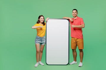 Full body smiling fun young couple two friend family man woman wear t-shirts together point finger on big huge blank screen mobile cell phone with area isolated on pastel plain light green background