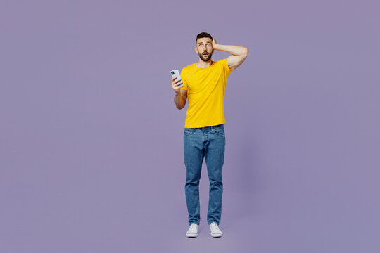 Full body young sad shocked fearful caucasian man wear yellow t-shirt use mobile cell phone hold head look camera isolated on plain pastel light purple background studio portrait. Lifestyle concept.