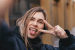 © zvkate - Stylish girl with blonde hair making selfie outside. Cool woman in fashion bomber showing peace sign, winking eye and smiling outdoors.