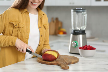 Wall Mural - Woman preparing mango for tasty smoothie at white marble table in kitchen, closeup