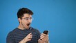 © Robert Peak - Surprised young man 20s posing isolated on blue background studio. Using mobile cell phone typing sms message say wow pointing.