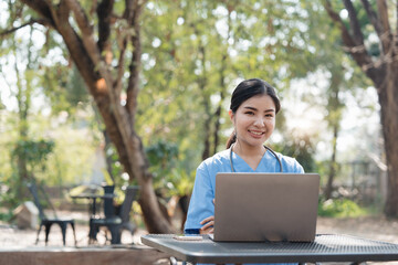 Wall Mural - Asian female nurse working outdoor video call with patient online on laptop at desk in park.